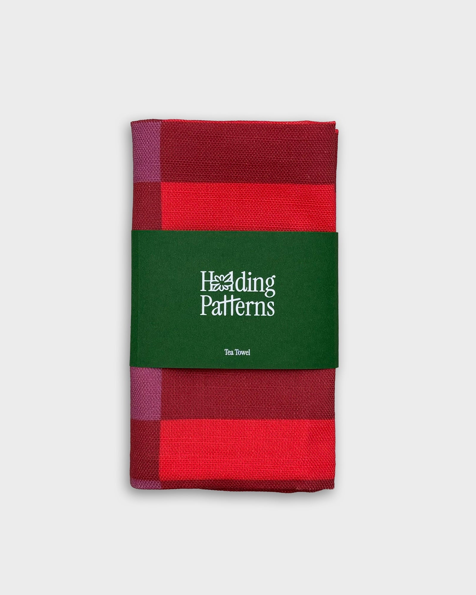 A folded tea towel with a red and magenta plaid pattern, wrapped in a dark green paper band that reads 'Hedding Patterns Tea Towel'.
